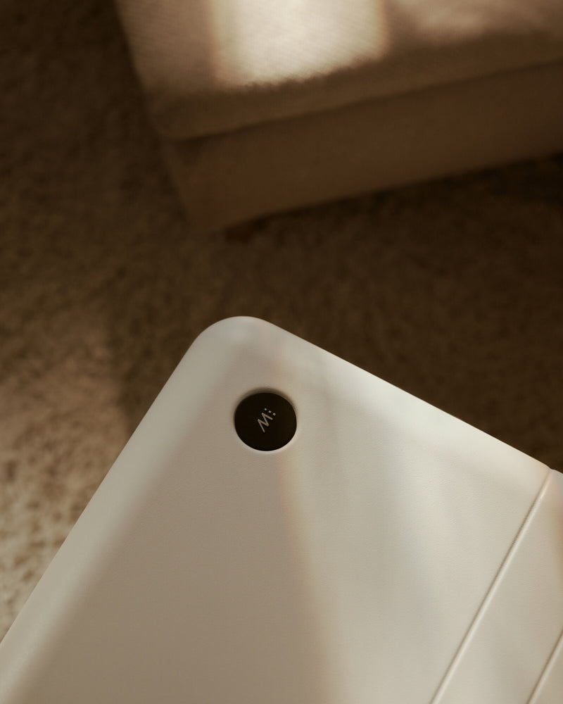 Close-up of a white appliance corner with a small circular black control button, photographed in warm indoor light with a carpeted floor and furniture blurred in the background.