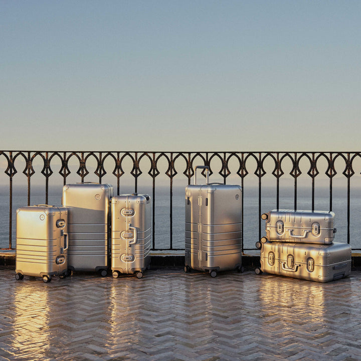 A collection of Monos aluminum suitcases in Aspen Silver in various sizes is arranged on a sunlit terrace with a wrought iron railing, overlooking the ocean in the background.