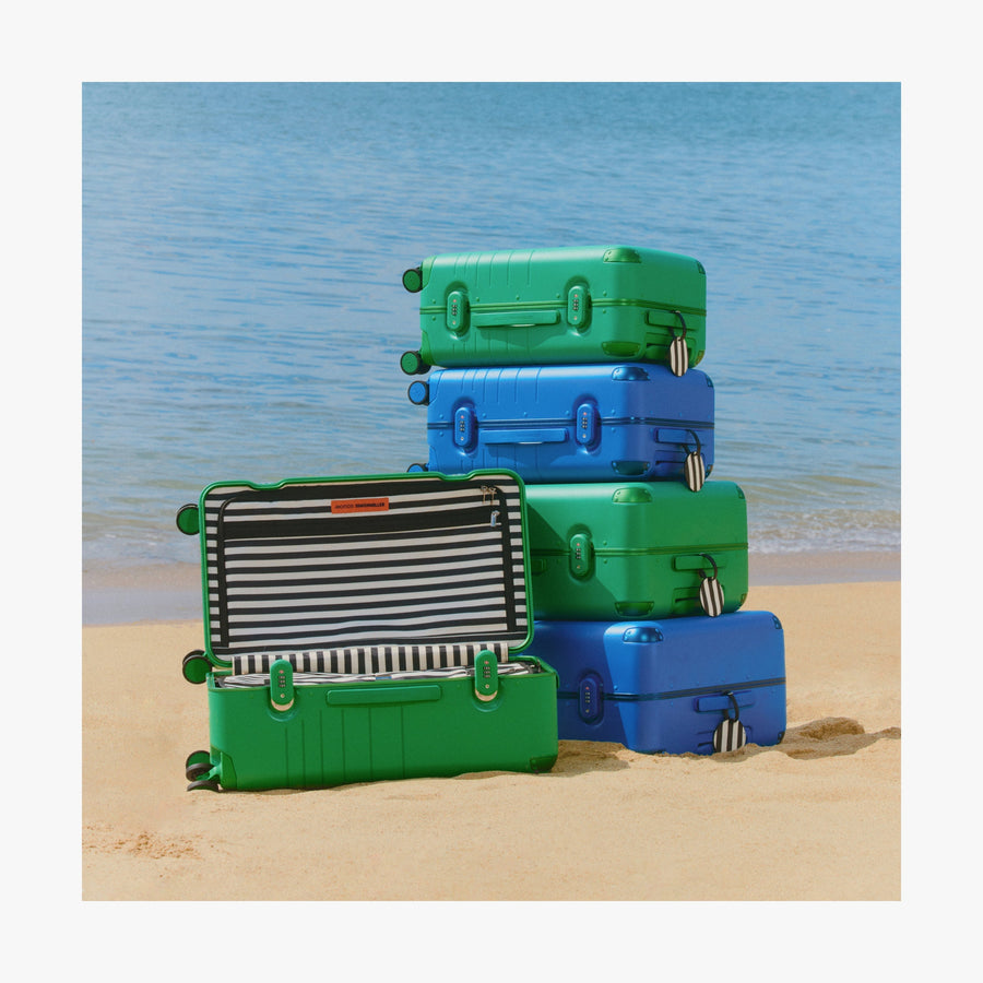 Coastal Blue | Stack of Jungle Green and Coastal Blue Hybrid suitcases on the sand, with one open suitcase revealing its striped interior