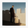 Adrien Brody wearing a dark coat and red scarf stands beside a white fortress-like wall, with a Monos aluminum carry-on plus in aspen silver in the sunlight. The deep blue sky contrasts with the shadows. (thumbnail)