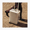 A close-up of a Monos aluminum carry-on plus suitcase in aspen silver. A person wearing dark brown trousers and polished black shoes walks alongside it on a textured stone pavement, with warm sunlight casting soft shadows. (thumbnail)