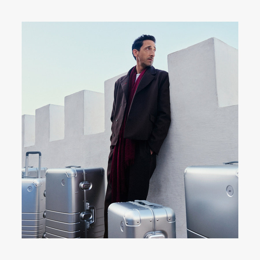 Aspen Silver | Adrien Brody leans against a white fortress-like wall, gazing into the distance, with multiple Monos aluminum suitcases in aspen silver around him.