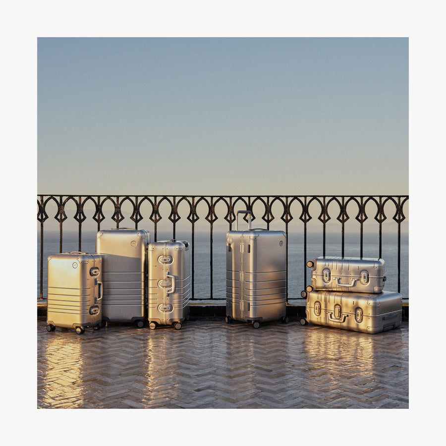 Caviar Black | A collection of Monos aluminum suitcases in Aspen Silver in various sizes is arranged on a sunlit terrace with a wrought iron railing, overlooking the ocean in the background.