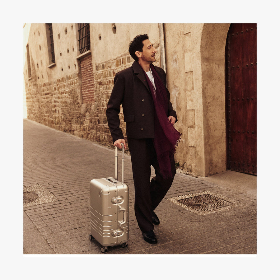 Caviar Black | Adrien Brody dressed in a dark brown suit, white shirt, and a deep burgundy scarf walks down a historic cobblestone street while pulling a Monos Aluminum carry-on plus suitcase in aspen silver. The background features an old stone building with a wooden door, adding to the image's timeless, travel-inspired aesthetic.