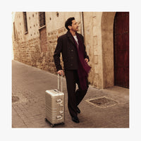 Adrien Brody dressed in a dark brown suit, white shirt, and a deep burgundy scarf walks down a historic cobblestone street while pulling a Monos Aluminum carry-on plus suitcase in aspen silver. The background features an old stone building with a wooden door, adding to the image's timeless, travel-inspired aesthetic. (thumbnail)