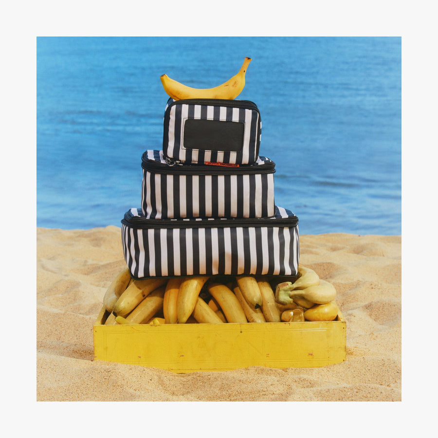 Set of Six / Poplin Stripe Black | Stack of Poplin Stripe Black compressible packing cubes arranged on a crate of bananas on a sandy beach, with a single banana placed on top.