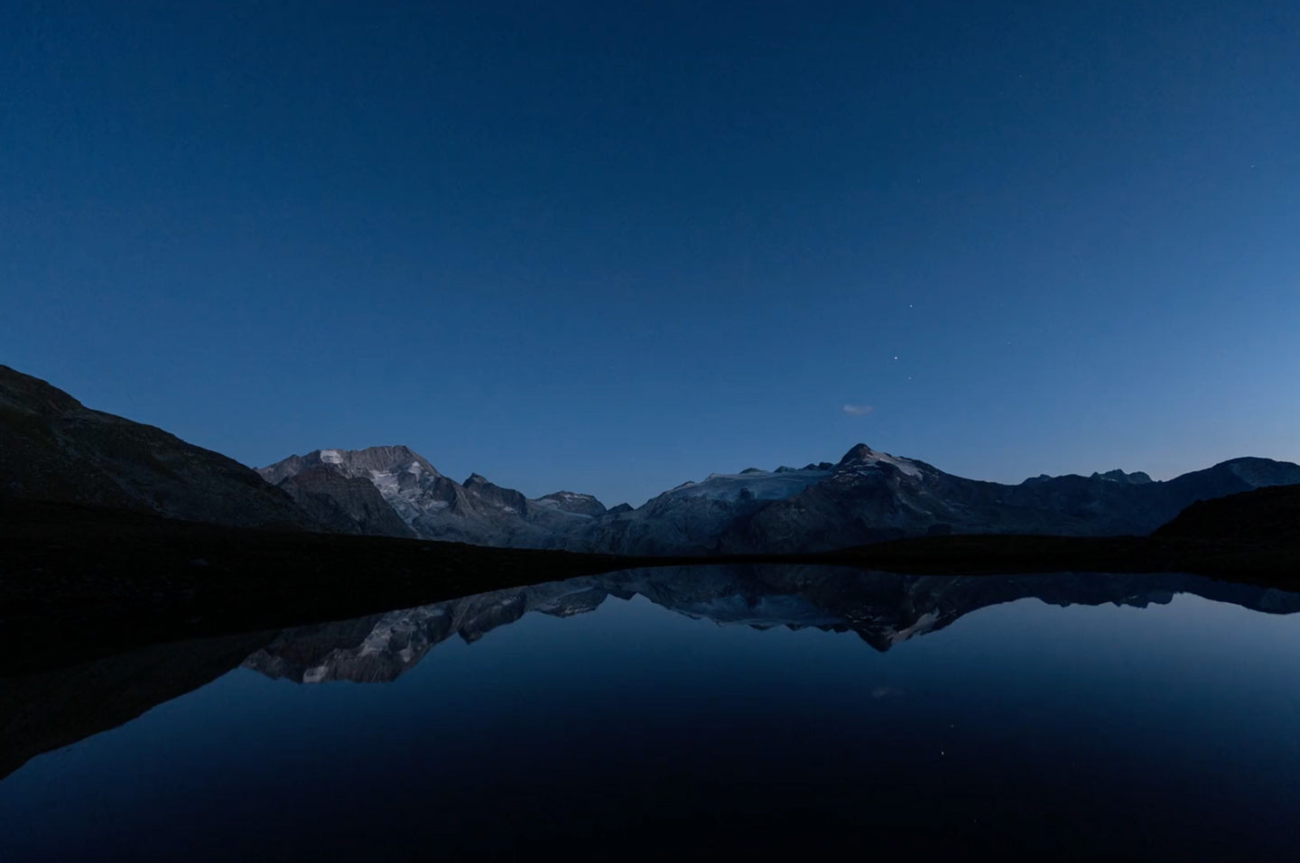 A serene mountain landscape at dusk, with snow-capped peaks reflecting on a still, glass-like lake under a deep blue sky with faint stars. The rugged slopes frame the tranquil water, creating a peaceful and symmetrical scene.