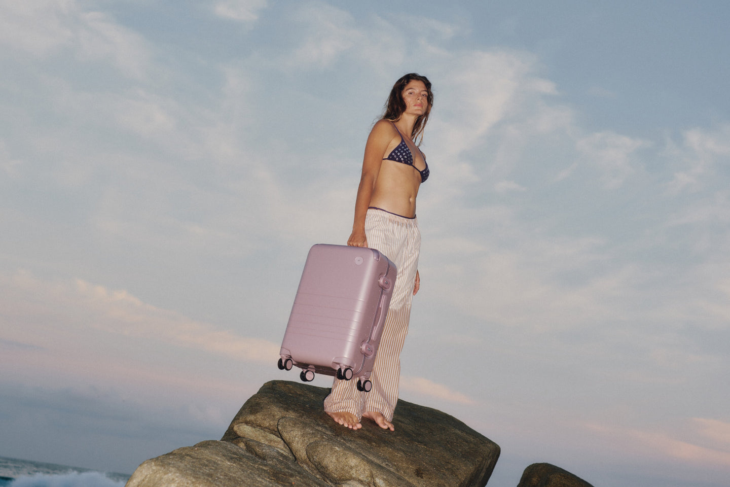A woman stands barefoot on a seaside rock holding the Monos hybrid Carry-On in Orchid, with the ocean and pastel sky behind her — capturing the spirit of travel and effortless elegance.
