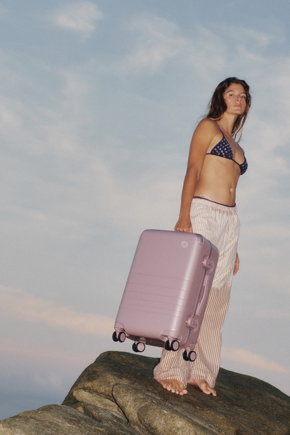 A woman stands barefoot on a seaside rock holding the Monos hybrid Carry-On in Orchid, with the ocean and pastel sky behind her — capturing the spirit of travel and effortless elegance.