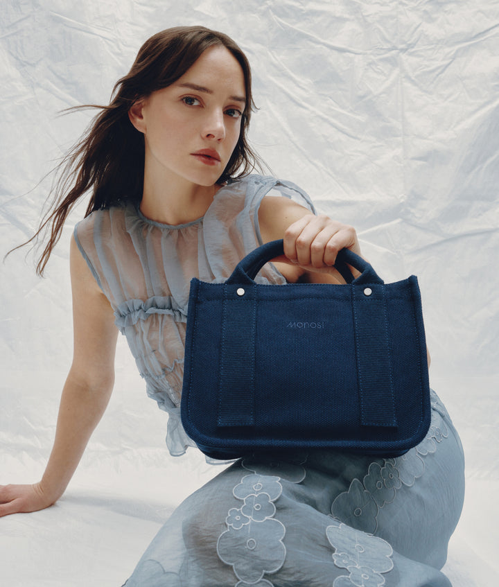 A woman holding the Monos Gesso Small Tote in Abyss Blue. The structured canvas tote is styled against a soft white backdrop, highlighting its minimalist design and refined texture.