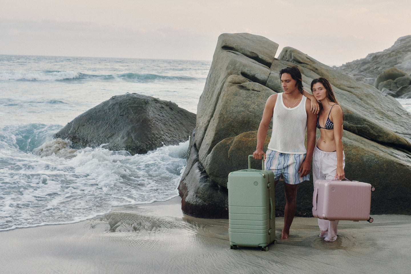 Two travelers stand barefoot at the shoreline with the Hybrid Trunk in Mint and the Hybrid Carry-On in Orchid, surrounded by ocean waves and coastal rock formations.
