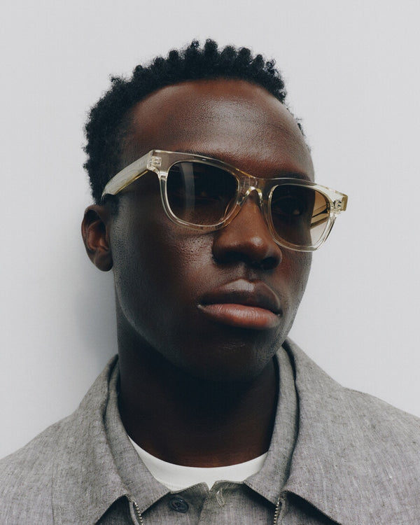 Man wearing gold-framed sunglasses and a gray coat against a white background