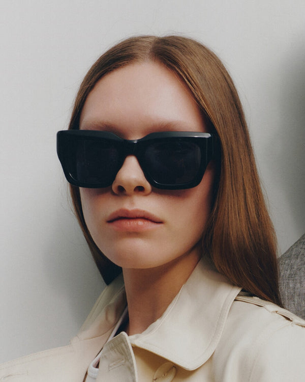 Woman wearing black sunglasses and a beige coat against a neutral background
