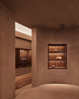 Curved hallway inside the Monos SoHo store featuring recessed wood shelving with neatly displayed travel bags and aluminum luggage, illuminated by warm ambient light against terracotta brick walls.