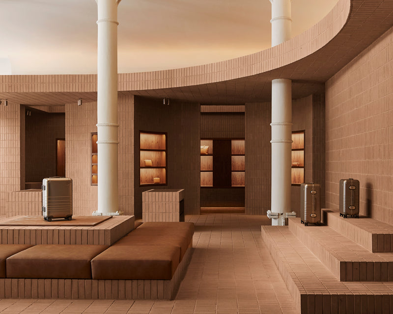 A modern architectural interior with warm terracotta brick walls and columns, brown upholstered seating, built-in shelving, and two metallic suitcase-like objects placed on raised platforms.