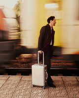 Adrien Brody walks along a train platform with a Monos aluminum carry-on plus in aspen silver, as a yellow train rushes past in motion blur.