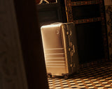 A Monos aluminum trunk in aspen silver stands alone inside a dimly lit room with patterned tile flooring, partially framed by a dark wooden door.