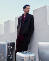 Adrien Brody leans against a white fortress-like wall, gazing into the distance, with multiple Monos aluminum suitcases in aspen silver around him.